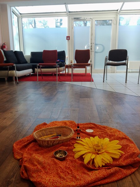 Blick in den Seminarraum. Vorn: Rote Decke auf dem Boden mit gelber Blüte. Hinten: Stuhlkreis vor Fensterfront